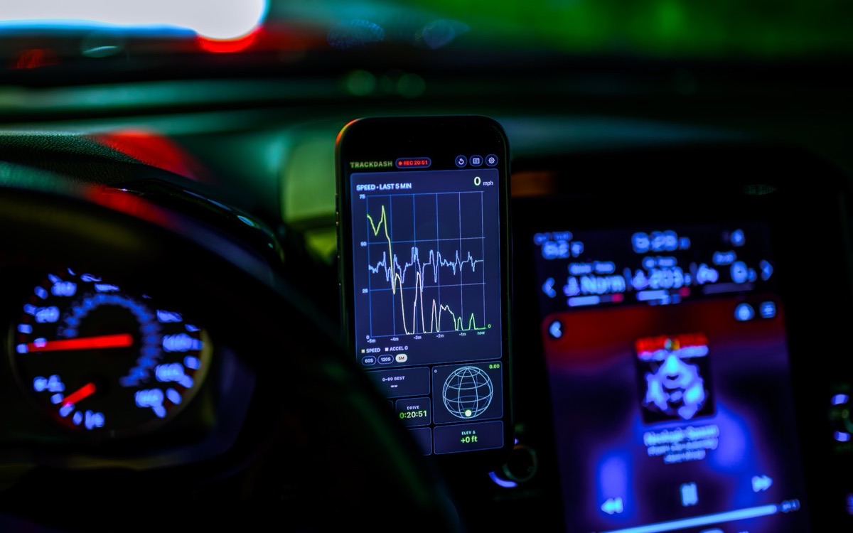 TrackDash running on a phone mounted in a car at night, showing a five-minute speed trace beside the instrument cluster and the CarPlay screen.